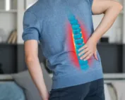 A person is holding their Herniated Discs, overlaid with a graphic indicating spinal pain, wearing a grey shirt and jeans.
