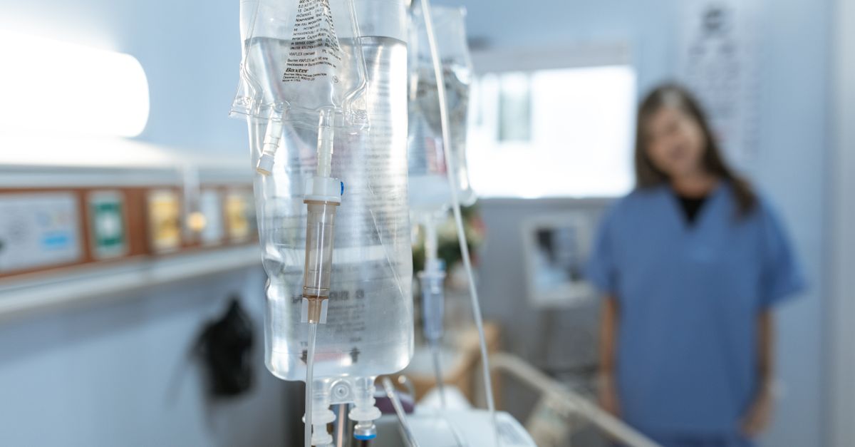 Close-up of an IV drip in a hospital with a blurred nurse in the background.