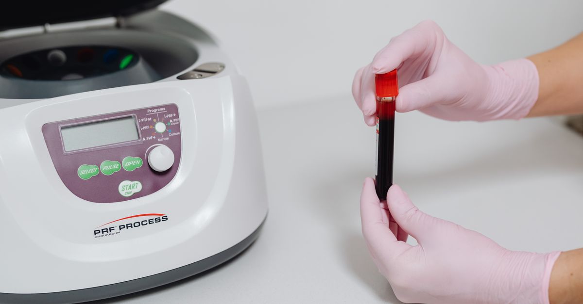 Close-up of hands in gloves holding a blood sample beside a centrifuge.